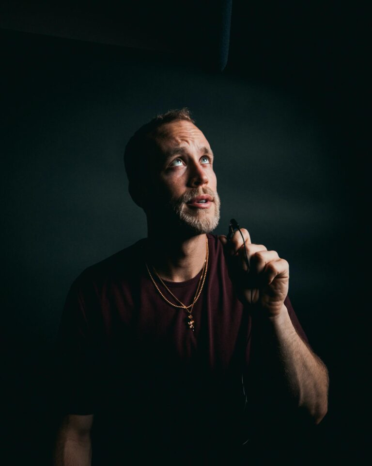 Medium close-up portrait of a thoughtful man with a beard holding a microphone under studio lighting.